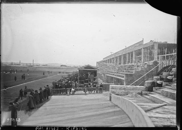 Le stade de Gerland en construction : une foule assiste à un match devant une tribune de béton inachevée, entourée d'échafaudages.
