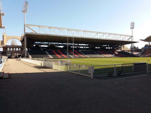 L'intérieur du stade de Gerland aujourd'hui, tribune couverte aux sièges noirs et rouges, avec une arche en béton visible sur la gauche.